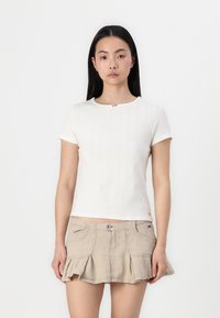 Hvit kortermet topp med teksturert mønster, rund hals og en liten accentknapp. Kombinert med en beige, rynket kort skjørt.