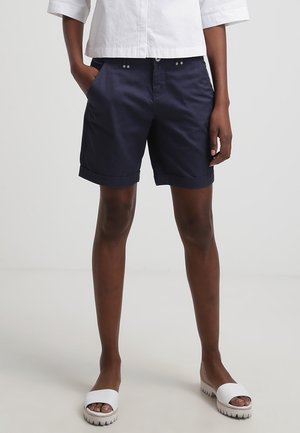Personne portant un short marine à ourlet retroussé, une chemise blanche raccourcie et des sandales blanches ouvertes aux orteils, debout avec une main dans la poche devant un fond uni.