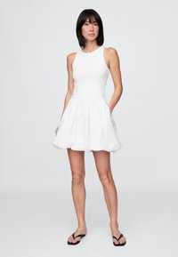 Biele šaty bez rukávov s priliehavým živôtikom a rozšírenou, textúrovanou sukňou. Výrazné sú okrúhly výstrih a bočné vrecká.