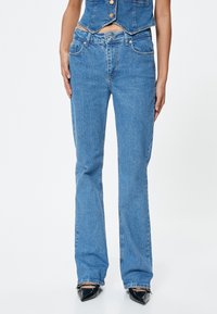 Jeans de mezclilla azul de corte acampanado con una cintura de altura media, cinco bolsillos, ligeros desgastes y sutiles detalles de arrugado. Textura suave con un ajuste entallado.