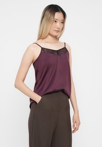 Camisola boje vina od glatkog materijala, s dekolteom ukrašenim šljokicama. Podesive naramenice i opušteni kroj.