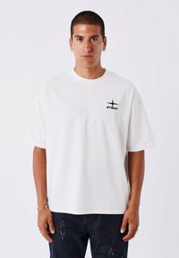 Camiseta de algodón blanca de manga corta, con cuello redondo, que presenta un pequeño logo negro y un diseño de estrella en la parte izquierda del pecho.