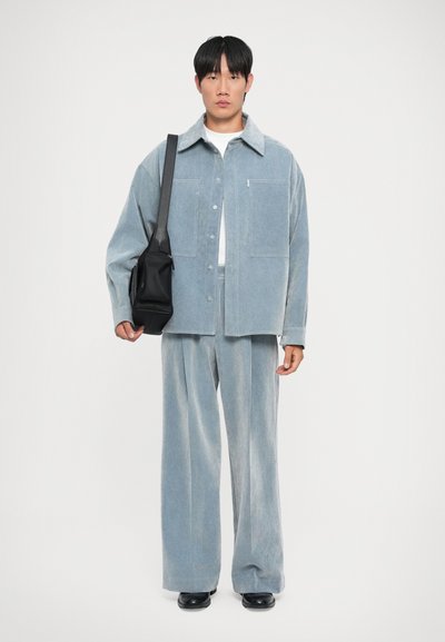 Hellblaues Cordjackett mit schrägen Taschen, kombiniert mit passenden weit geschnittenen Hosen. Das Model hält eine schwarze Umhängetasche.