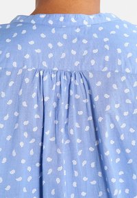 Tissu de chemise bleu clair avec un motif paisley blanc, présentant un empiècement plissé au dos et un col arrondi. Texture douce, design décontracté.