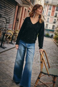 Juodas V formos kaklo megztinis su blizgančiomis detalėmis, derinamas su plačiomis, išblukusiomis mėlynomis džinsinėmis kelnėmis. Laiko bistro kėdę su pintu sėdimuoju.