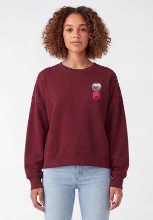 Jeune femme aux cheveux bouclés portant un sweat-shirt bordeaux avec un petit graphisme coloré de distributeur de chewing-gum sur le côté gauche de la poitrine.