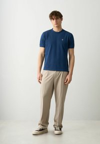 Mörkblå kortärmad t-shirt med rund halsringning, mjukt bomullsmaterial och en liten logotyp på bröstet, kombinerat med beige byxor och sneakers.