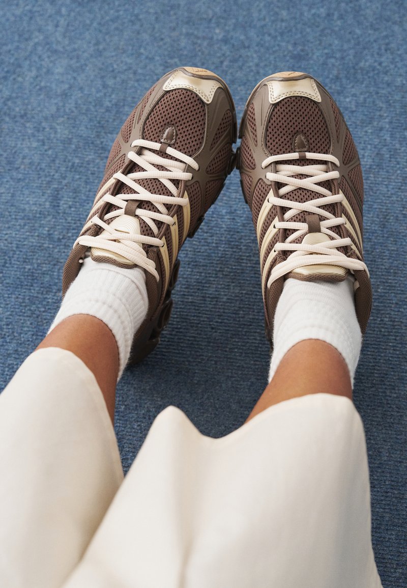 Ruskeat urheilukengät, joissa valkoiset nauhat, verkkokuosi, beigejä yksityiskohtia ja pyöristetty kärki, yhdistetty valkoisiin resoraisiin sukkiin.