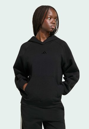 Jonge vrouw met gevlochten haar, een zwarte Adidas hoodie en bijpassende broek, handen in de zakken, kijkt opzij tegen een lichte achtergrond.