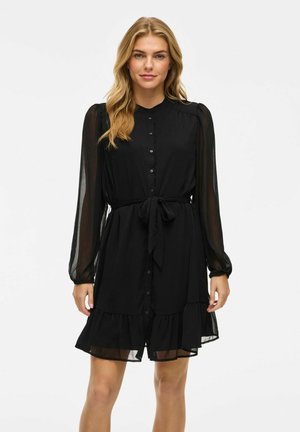 Vestito nero con chiusura a bottoni, maniche lunghe trasparenti, vita con nastro da annodare e orlo a balze a strati. Tessuto morbido e leggero.