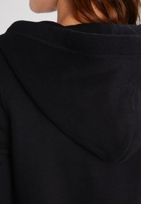 Svart huvtröja med mjukt, texturerat tyg, ribbat halsband och en djup huva. Baksidan har minimala sömnadsdetaljer.