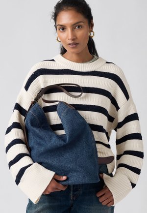 Γυναίκα με σκούρα μαλλιά φοράει πουλόβερ με κρεμ και μαύρες ρίγες και μπλε τζιν, κρατώντας μεγάλη τσάντα ώμου από τζιν με καφέ λουρί.