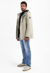 Lichtbeige waterdichte jas met een capuchon, ritssluiting aan de voorkant en zijzakken. Ggedragen over een donkere top, gecombineerd met een blauwe jeans en grijze sneakers.