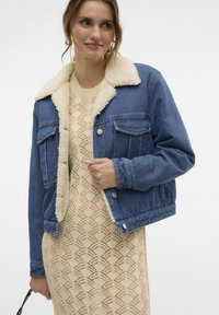 Veste en denim avec un col en fausse fourrure beige doux, dotée de deux poches poitrine, d'une fermeture à boutons et d'un design raccourci. Portée sur une robe en tricot.