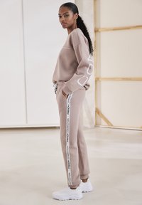 Beige sweatshirt met witte grafische print op de mouw, gecombineerd met beige joggers met zijstrepen. Witte sportschoenen maken de outfit compleet.