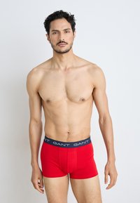 Červené pánske boxerky s námorníckym pásom, na ktorom je názov značky "GANT" v bielom texte. Tkanina vyzerá hladko a je priliehavá.