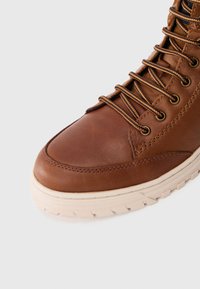 Smeđi kožni čizmi s okruglim vrhom, svijetlosmeđim gumenim potplatom i kontrastnim crnim i smešnim vezicama. Ima metalne rupe za vezivanje i vidljive šavove.