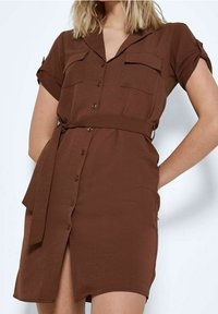 Hnědé šaty košilového střihu s krátkým rukávem, zapínáním na knoflíky vpředu, dvěma náprsními kapsami a vlastním opaskem. Hladká textura; detail s jedním bočním rozparkem.