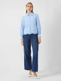 Light blue button-up shirt with eyelet detailing, long sleeves, and a scalloped hem, paired with high-waisted dark denim jeans and white heels.