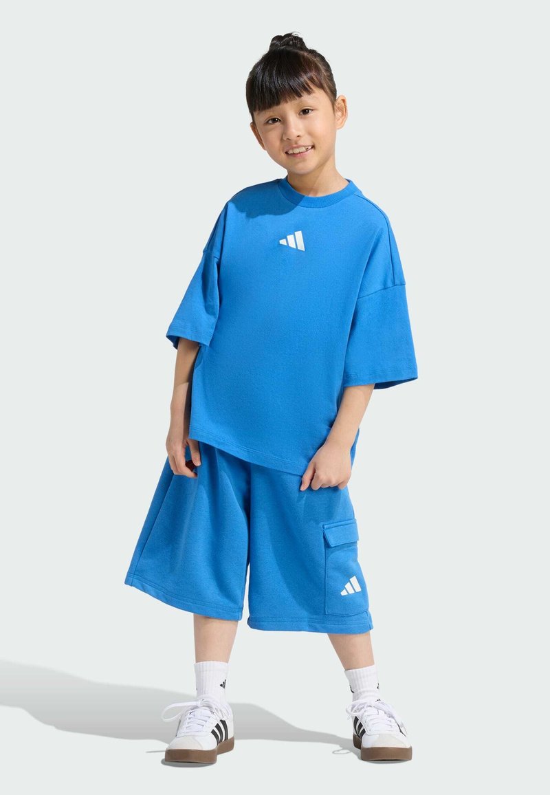 Modrý športový outfit, ktorý pozostáva z trička s krátkym rukávom a šortkami po kolená. Materiál vyzerá mäkko a na oboch kusoch sú akcenty loga Adidas.
