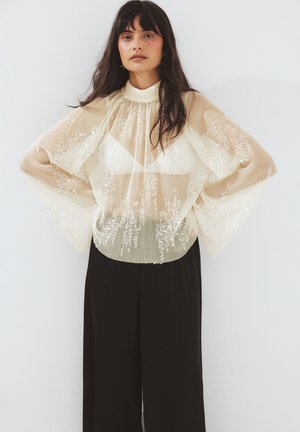 Przezroczysta beżowa bluzka z plisami i kwiatową haftem, z wysokim kołnierzem i szerokimi rękawami; zestawiona z czarnymi spodniami o szerokich nogawkach.