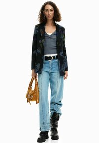 Blazer nero con motivo floreale sopra una maglia grigia, abbinato a jeans azzurri a vita alta e stivali neri al ginocchio, con una borsa color senape in mano.