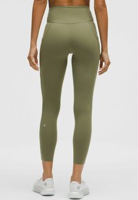 Hochbundige, olivgrüne Leggings aus dehnbarem, glattem Material mit Seitentaschen und einer figurbetonten Passform, kombiniert mit weißen Sportschuhen.