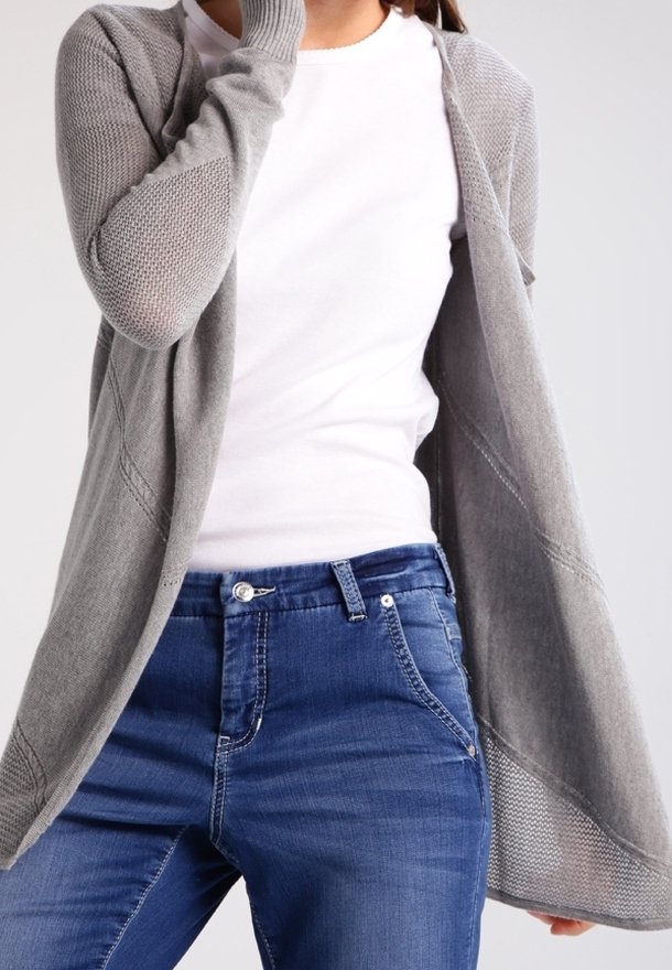 Femme portant un t-shirt blanc, un jean bleu et un cardigan en maille gris ample, une main près du visage.