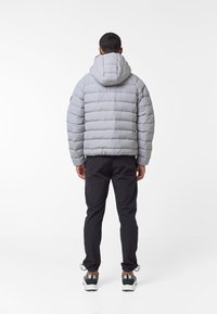Giacca imbottita grigia con cappuccio, caratterizzata da cuciture orizzontali e polsini elasticizzati. Indossata sopra pantaloni neri e sneakers grigie. Sfondo bianco.