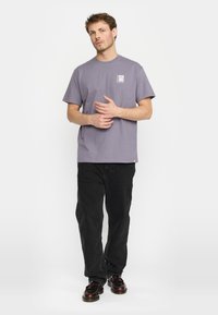 Paarse katoenen T-shirt met korte mouwen, ronde hals en kleine grafische print op de linker borst; gedragen met zwarte jeans en schoenen.