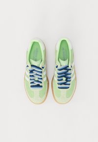 Gröna mockasneakers har vita ränder, tjocka blåa och vita mönstrade snören samt en beige gummisul och en grön innersula.