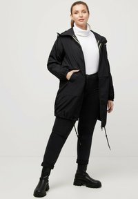 Svart hooded jacka med elastiska ärmslut, framfickor och dragsko i midjan. Bärs över en vit polotröja och kombineras med svarta jeans och stövlar.