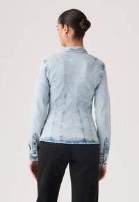 Γυναίκα με μαλλιά σε κότσο, φοράει εφαρμοστό ανοιχτό γαλάζιο τζιν μπουφάν και μαύρο παντελόνι, στέκεται με την πλάτη γυρισμένη σε απλό φόντο.