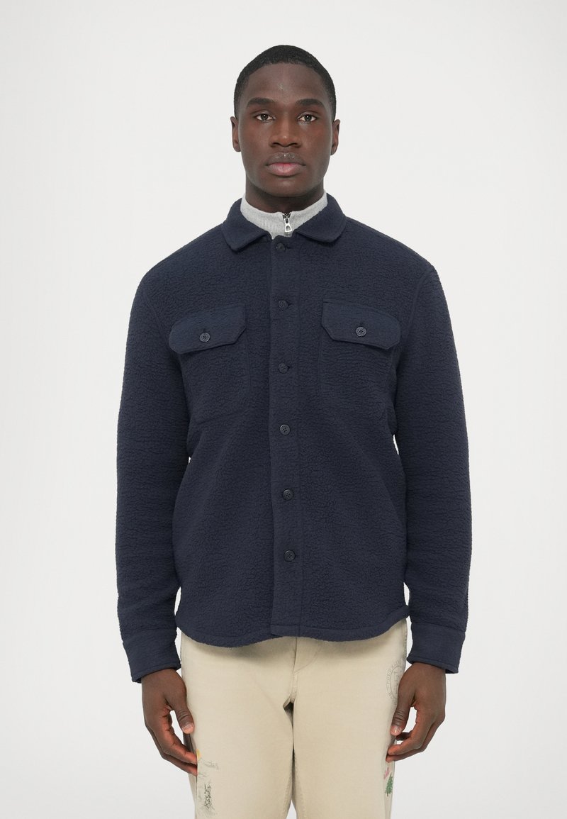 Mornarska majica-jakna iz flisa z teksturirano površino, dvema sprednjima žepoma, zapenjanjem na gumbe in ovratnikom. Nošena čez sivi pulover z zadrgo.