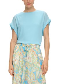 Blouse légère bleu clair à manches courtes en tissu transparent, associée à une jupe à motifs bleue, jaune et beige, avec une taille à cordon.
