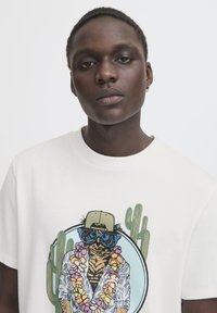White t-shirt featuring a colorful graphic of an animal in sunglasses and a floral lei, with cacti illustrated in the background.