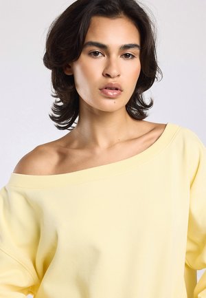 Vrouw met kort donker haar, dragend een losse lichtgele off-shoulder top, neutrale uitdrukking, tegen een effen lichtgrijze achtergrond.