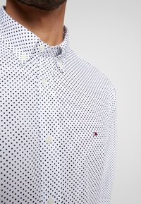 Witte button-downshirt met een marineblauw stippenpatroon, zichtbare knopen en een klein geborduurd logo in rood en blauw nabij de borst.