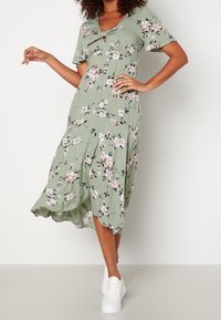 Frau in einem grünen Blumenkleid mit kurzen Ärmeln und weißen Sneakers, steht vor einem schlichten Hintergrund.