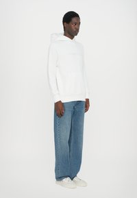 White hooded sweatshirt with front pocket and subtle branding, paired with loose-fitting blue denim jeans and white sneakers.