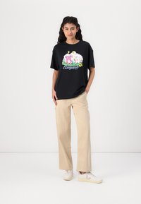 Schwarzes, oversized T-Shirt mit einem bunten Cartoon-Druck mit Tieren, kombiniert mit hellbeigen Cordhosen und weißen Sneakers.
