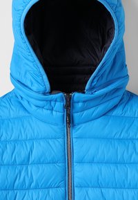 Modrá zateplená bunda s vysokým límcem a kapucí. Má vertikální prošité linie a centrální zip s jezdcem. Vnitřní podšívka je tmavá.