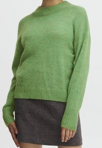 Green wool sweater with a ribbed round neckline and long sleeves. Soft texture, relaxed fit, displayed over a dark gray mini skirt.
