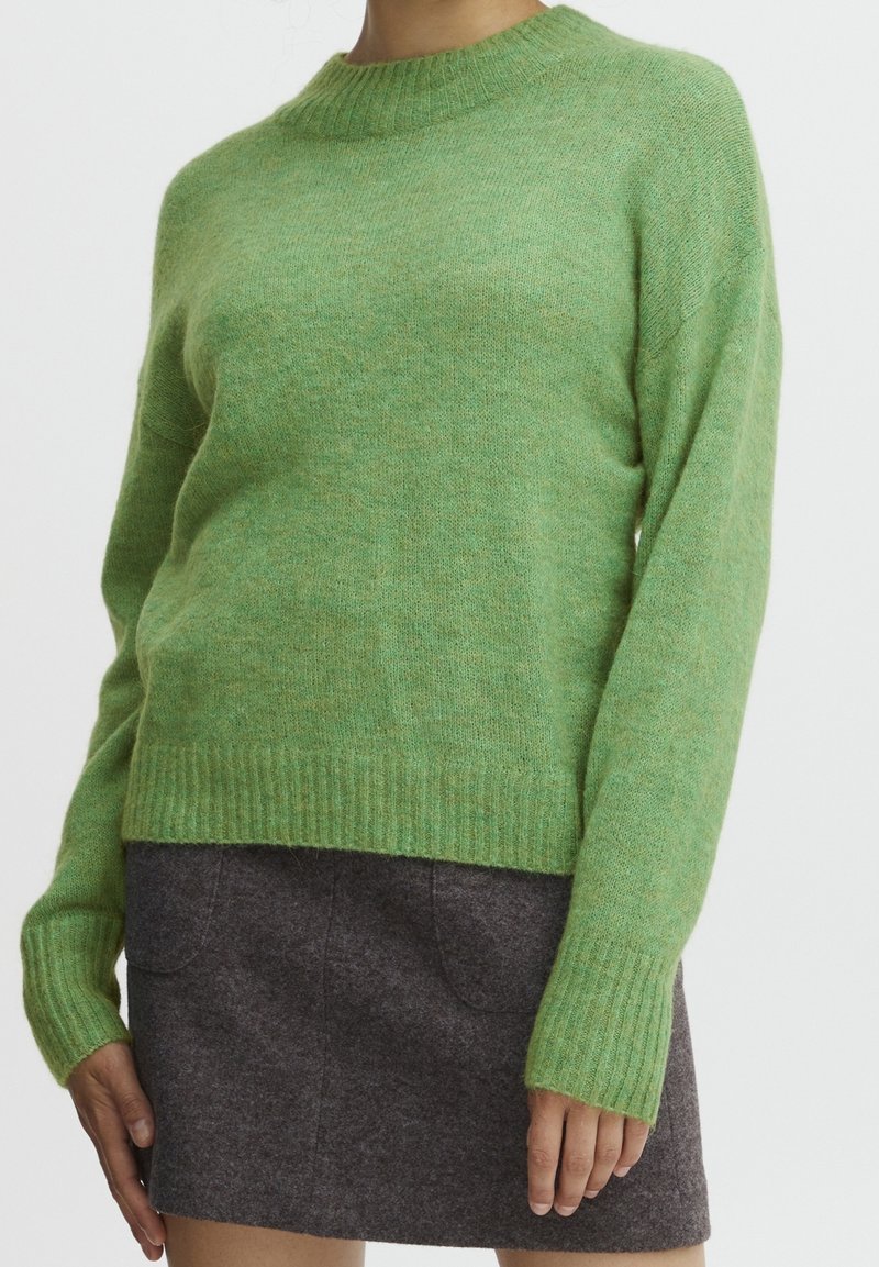 Green wool sweater with a ribbed round neckline and long sleeves. Soft texture, relaxed fit, displayed over a dark gray mini skirt.