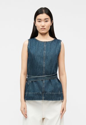 Joven mujer con un top de mezclilla sin mangas y un cinturón abotonado a un lado, combinados con pantalones blancos holgados, posando frente a un fondo liso.