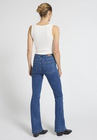 Frau mit Hochsteckfrisur, die ein weißes ärmelloses Oberteil, blaue Schlagjeans und schwarze Stiefeletten mit Absatz trägt, steht mit dem Rücken zur Kamera.