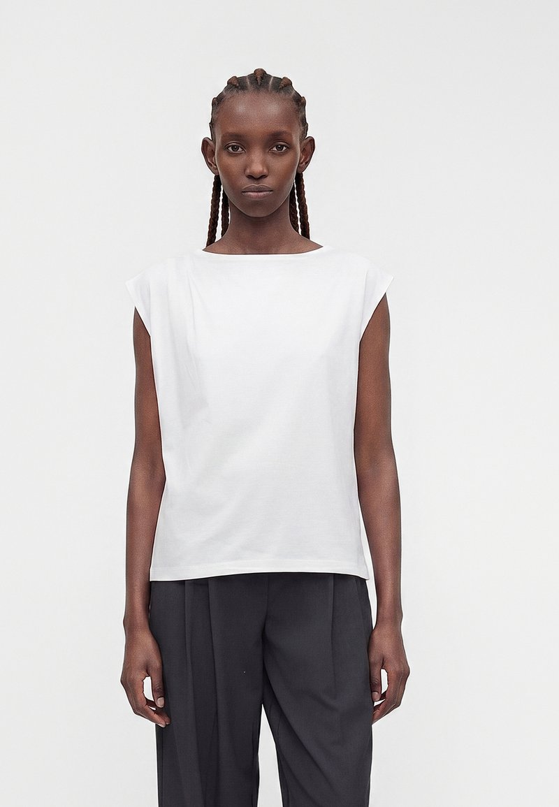 Femme aux cheveux tressés, debout face à la caméra, portant un haut blanc sans manches et un pantalon noir plissé sur un fond uni.