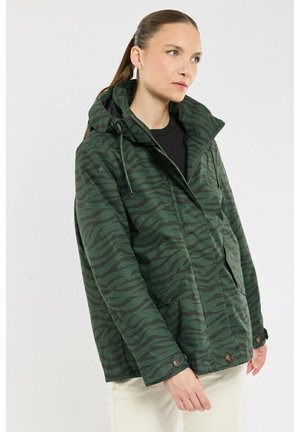 Femme portant une veste à capuche vert et noir à rayures zébrées par-dessus un haut noir et un pantalon blanc sur un fond clair uni.