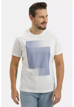 Mężczyzna z brodą, ubrany w biały T-shirt z pionowym niebieskim paskowanym wzorem, jasnoniebieskie dżinsy, patrzy w prawo, uśmiecha się na jednolitym tle.