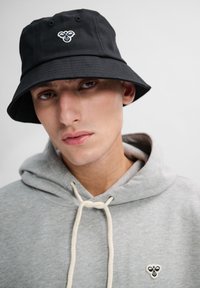 Svart bucket-hatt i bomull, med en dekorativ broderad logotyp framtill. Buren över en grå hoodie med snörning.